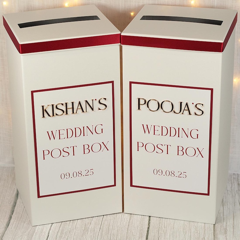 May include: Two tall, rectangular white wedding post boxes with a horizontal slot at the top. Each box features a dark red border and text reading KISHAN'S WEDDING POST BOX 09.08.25 and POOJA'S WEDDING POST BOX 09.08.25 in dark red and gold lettering.