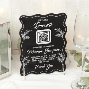 May include: A black acrylic sign with a decorative wavy edge. The sign reads "PLEASE Donate" above a QR code, with text below stating "In Loving Memory of Marion Simpson." Additional text encourages donations to a charity.