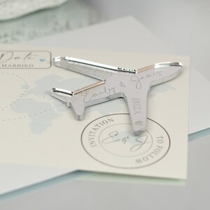 May include: Silver airplane shaped wedding invitation with the text "Emily & James" and "IBIZA" on the plane. The invitation also has a circular stamp with the text "INVITATION" and "E&J" in cursive.