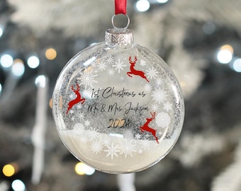 Könnte beinhalten: Klare Glas-Weihnachtskugel mit einem weißen Schneeflockenmuster und roten Rentier-Silhouetten. Die Kugel enthält eine weiße, schneeähnliche Substanz und den Text "1. Weihnachten als Herr und Frau Jackson 2021".