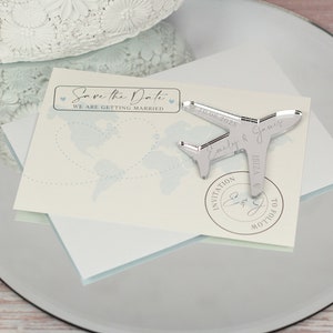 May include: Save the Date card with a silver airplane charm. The card features a world map with a blue dotted line and the text "Save the Date" with hearts. The charm has the text "Emily & James 10.08.2025 IBIZA" and a stamp that says "Invitation to follow".