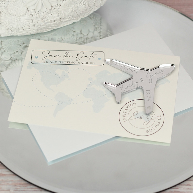 May include: Save the Date card with a silver airplane charm. The card features a world map with a blue dotted line and the text "Save the Date" with hearts. The charm has the text "Emily & James 10.08.2025 IBIZA" and a stamp that says "Invitation to follow".