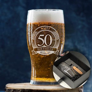 May include: A pint glass filled with beer and a white head, engraved with "Happy 50th Birthday Robert." The glass is on a wooden surface. A gift box contains a matching glass and bottle opener.