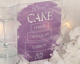 May include: A clear acrylic sign with the words "Let's eat CAKE" in silver lettering. Below, the sign lists cake flavors: Lemon, Chocolate, and Carrot. The sign has a purple background with white accents and the initials "A S" and the date 23.08.25.
