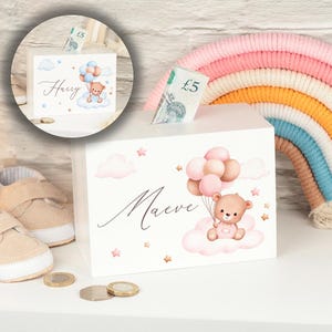 May include: White wooden money box with a pink and white cloud design. A teddy bear is holding pink and white balloons. The name "Maeve" is written in cursive on the box.