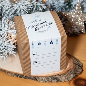 以下が含まれることがあります： 茶色の段ボール箱に、青いクリスマスオーナメントと雪の結晶が描かれた白い紙の帯が巻かれています。テキストには「A SPECIAL Christmas Keepsake HANDMADE BY Love June」と書かれており、「To」と「From」の記入欄があります。