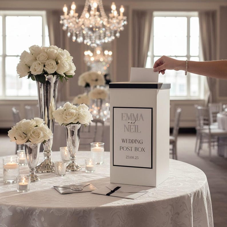May include: A white rectangular wedding post box with a black lid and a black-bordered label featuring the text EMMA and NEIL, WEDDING POST BOX, and 23.08.25. The box is placed on a white tablecloth alongside silver vases with white roses.