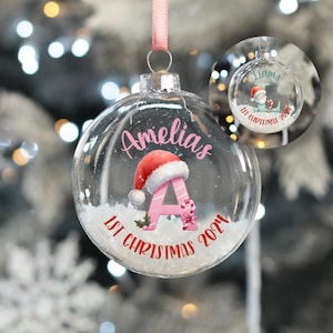 May include: A clear glass Christmas bauble with a pink ribbon and a pink letter A with a Santa hat on top. The bauble also has the text "Amelia's 1st Christmas 2021" on it.