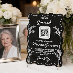 May include: A black memorial sign with white text that reads "Please Donate" and "In Loving Memory of Marion Simpson." It includes a QR code and the words "Thank You." The sign is displayed on a table with a framed photo and flowers.