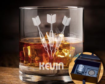 May include: A personalized whiskey glass with a design of three darts and the name "Kevin" etched on the bottom. The glass is filled with amber liquid and ice cubes. A small box with a dartboard and a miniature glass is visible in the background.