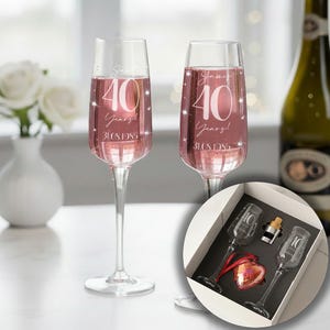 May include: Two clear glass champagne flutes filled with a pink liquid, each featuring white text that reads "James 40 Years! 31.08.1985" with star accents. A white gift box contains two identical flutes, a metal stopper, and a red heart ornament.