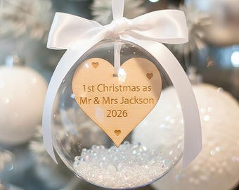 Könnte beinhalten: Eine klare, kugelförmige Weihnachtskugel mit kleinen weißen Perlen und einem herzförmigen Holzeinsatz. Das Herz ist mit dem Text 1st Christmas as Mr & Mrs Jackson 2026 graviert und mit kleinen herzförmigen Ausschnitten versehen.