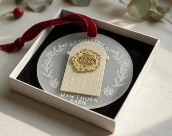 May include: A circular frosted acrylic ornament with a red velvet ribbon. It features a cream-colored door graphic, a gold wreath with the year 2026, and the text 1ST CHRISTMAS AT HAWTHORN BARN. The ornament is presented in a white gift box.