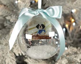 May include: A clear glass Christmas ornament with a photo of a dog inside. The ornament is decorated with faux snow and a blue ribbon. The text on the ornament reads "Harbie, The Brightest Star This Christmas."