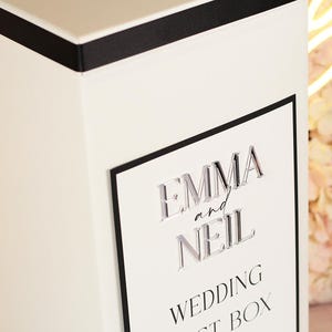 May include: A white rectangular box featuring a black border and a black top edge. The front displays the raised, clear acrylic text EMMA and NEIL in a serif font, followed by the printed black text WEDDING GUEST BOX in a simple sans-serif font.
