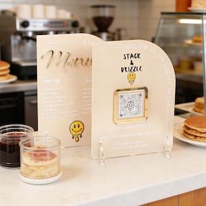 May include: Two cream acrylic signs, one a menu with gold lettering and a list of toppings, the other a social media sign with a QR code and the text "Stack & Drizzle". Jars of jam and pancakes are in the foreground. The signs are on a counter.