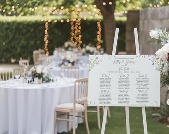 May include: A white easel displays a wedding seating chart with floral accents. The chart lists names under table numbers, with the names "Kelly & James" at the top. The background shows a garden setting with tables, chairs, and string lights.