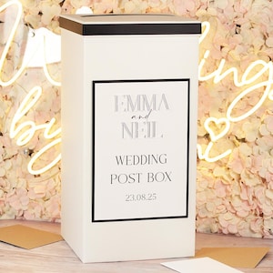 May include: A tall, rectangular white wedding post box with a black lid and a slot at the top. The front features a black-bordered label with the text EMMA and NEIL, WEDDING POST BOX, and the date 23.08.25 printed in a clean, black serif font.