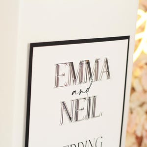 May include: A rectangular white sign with a thin black border featuring the raised, three-dimensional clear acrylic names EMMA and NEIL in a bold serif font, with the word and written in a smaller, black, cursive script between the names.