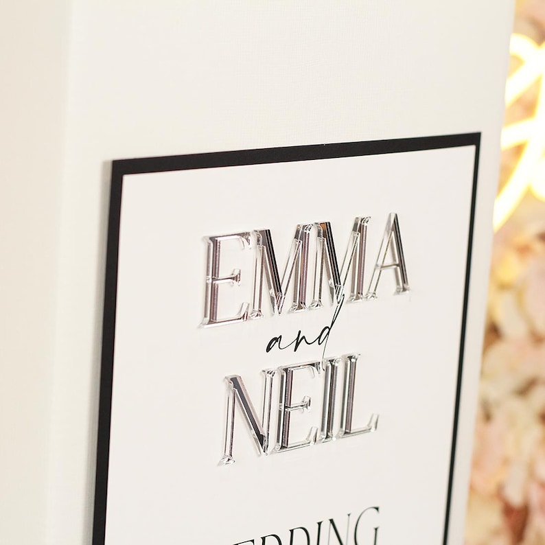 May include: A rectangular white sign with a thin black border featuring the raised, three-dimensional clear acrylic names EMMA and NEIL in a bold serif font, with the word and written in a smaller, black, cursive script between the names.