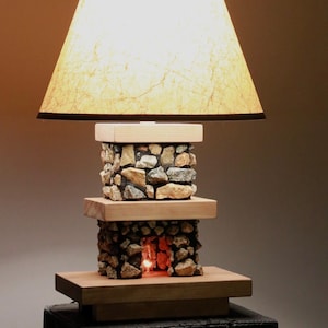 May include: A unique table lamp with a natural, rustic design. The lamp features a beige lampshade with a dark brown trim. The base is constructed with stacked stones and wooden platforms, creating a warm, inviting glow. The lamp is on a black stand.