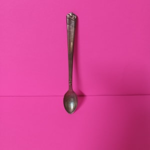 Puede incluir: Una cuchara de plata con un mango adornado sobre un fondo rosa brillante.