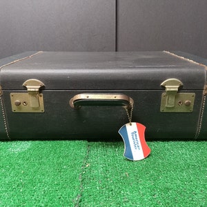 May include: A vintage black suitcase with brass hardware and a leather handle. The suitcase has a label that reads "American Tourister".