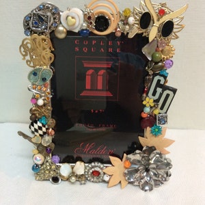 May include: A 5x7 inch photo frame decorated with various colorful jewels, buttons, and trinkets. The frame is made of metal and has a black background with the words "Copley Square" and "Malden International Designs" printed on it.