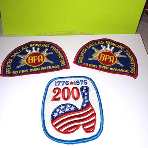 Könnte beinhalten: Drei gestickte Aufnäher. Zwei identische blaue halbkreisförmige Aufnäher zeigen den Text Greater Dallas Bowling Proprietors und 50 Pins Over Average mit einem BPA-Logo. Ein weißer rechteckiger Aufnäher zeigt 1776-1976, 200 und eine Bowlingkegel.