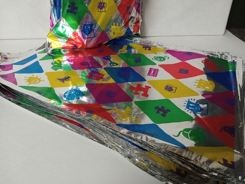Puede incluir: Una pila de bolsas de regalo de papel de aluminio plateado y coloridas con un patr&oacute;n repetitivo de varias formas y s&iacute;mbolos. Las bolsas est&aacute;n dobladas y apiladas unas sobre otras.