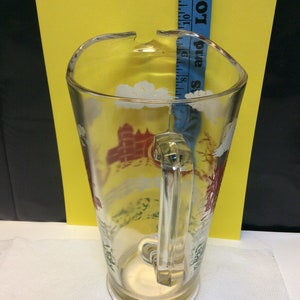 May include: A clear glass pitcher with a handle, featuring a painted design of a red and white castle with green trees and white clouds. The pitcher is on a white surface.