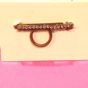 Può includere: Un anello in metallo color oro con una barra orizzontale fissata sulla parte superiore. La barra è incastonata con una fila di piccoli strass trasparenti e scintillanti che ne percorrono la lunghezza. L'anello è montato su un cartoncino bianco.