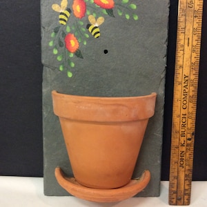 May include: A slate wall planter with a painted design of flowers and bees. The planter has a terracotta pot with a matching saucer.
