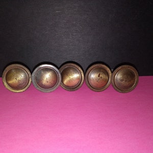 Può includere: Sei pomelli per armadi in ottone antico con forma arrotondata e bombata. I pomelli sono disposti in fila su una superficie rosa.