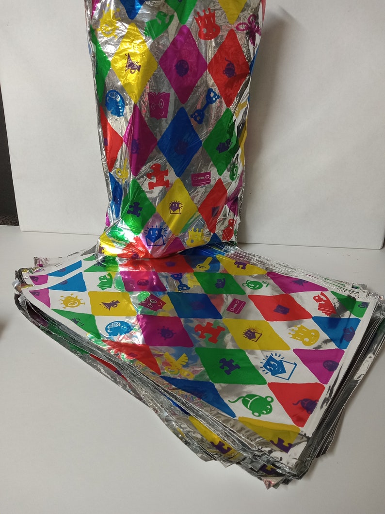Puede incluir: Una pila de hojas de papel de regalo de aluminio plateado y colorido con un patr&oacute;n de diamantes y varias im&aacute;genes de dibujos animados. El papel de regalo presenta una variedad de colores, incluyendo rojo, amarillo, azul, verde y morado.