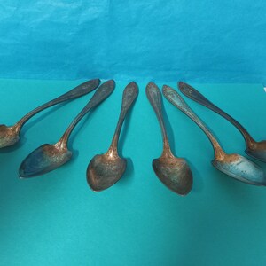 Puede incluir: Juego de seis cucharas plateadas con mangos ornamentados. Las cucharas tienen un acabado deslustrado y están dispuestas en forma de abanico sobre un fondo turquesa.