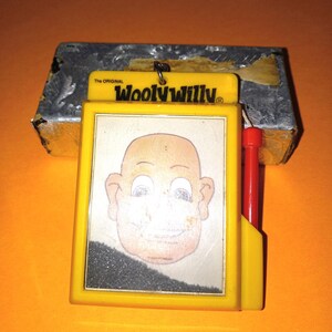 May include: The Original Wooly Willy magnetic drawing toy features a yellow plastic frame with a clear window displaying a cartoon face. A red magnetic wand is attached to the side by a string, used to move black magnetic filings to create hair.