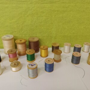 Op de afbeelding: Een verzameling houten spoelen met draad in verschillende kleuren, waaronder wit, bruin, groen, blauw en roze. De spoelen zijn gerangschikt op een wit oppervlak.