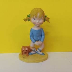 Könnte beinhalten: Eine Porzellanfigur einer jungen Ballerina in einem blauen Trikot und Ballettschuhen, die einen kleinen braunen Hund hält. Das Mädchen hat blonde Haare und einen Verband am Bein. Die Figur steht auf einem gelben Sockel mit der Signatur des Künstlers.