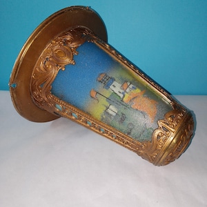 Może przedstawiać: Antyczny klosz w kolorze mosiądzu z ozdobnym, filigranowym wzorem. Klosz przedstawia malowaną scenę budynków i błękitne niebo. Klosz ma szeroki, okrągły wierzch i cylindryczny korpus.