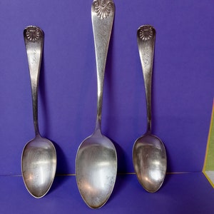 Pode incluir: Três colheres de prata com cabos ornamentados e letras gravadas. As colheres estão dispostas em fila sobre um fundo roxo.