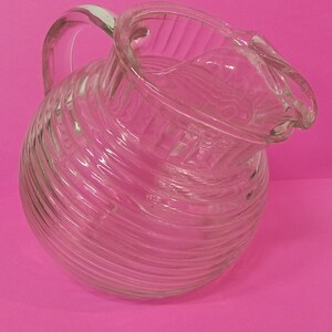 May include: A clear glass pitcher with a ribbed design and a handle. The pitcher is empty and has a slight tilt.