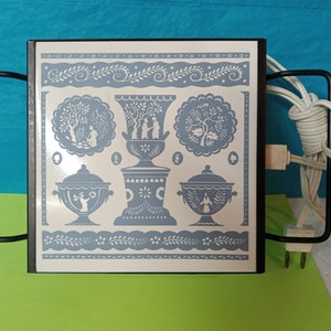Puede incluir: Una bandeja calentadora eléctrica blanca con marco negro y diseño floral azul y blanco. La bandeja tiene dos asas y un cable.