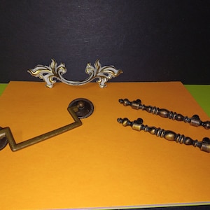 Op de afbeelding: Drie antieke kastbeslagknoppen. Eén is een sierlijke witte en gouden knop met een gebogen vorm. De andere twee zijn messing knoppen met een eenvoudig ontwerp.