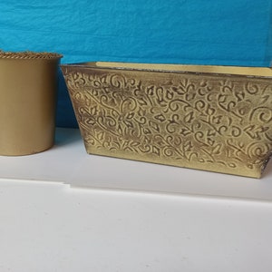 May include: Two gold-colored metal planters. The smaller planter is round and the larger planter is rectangular with a decorative embossed floral pattern.