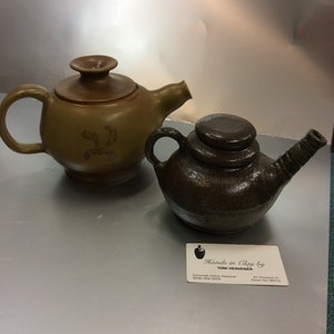Puede incluir: Dos teteras de cerámica, una marrón con tapa y la otra marrón oscuro con tapa de varias capas. Ambas tienen un pico y un asa. Las teteras están sobre una superficie plateada. Una tarjeta de visita para "Hands in Clay by Toni Heavener" está en la esquina inferior derecha.