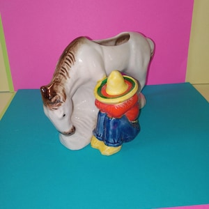 May include: A ceramic planter in the shape of a white horse with a brown mane and a small figure of a person wearing a sombrero, red jacket, blue pants, and yellow boots. The planter is on a blue surface with a pink background.