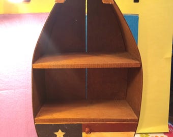 May include: A boat-shaped wooden wall shelf with two open storage levels and a bottom drawer. The drawer front features a painted design of a black square with a yellow star and horizontal red and yellow stripes resembling a flag.
