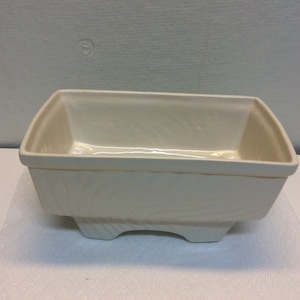 Puede incluir: Un macetero de cerámica blanco con forma rectangular y un diseño texturizado. El macetero tiene cuatro pequeñas patas.