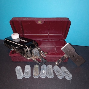 Może przedstawiać: Zestaw akcesoriów do maszyny do szycia Singer w stylu vintage. Zestaw zawiera czarno-srebrną maszynę do szycia, bordową walizkę, srebrny metalowy element i kilka szarych plastikowych nasadek. Widoczne jest logo Singer.